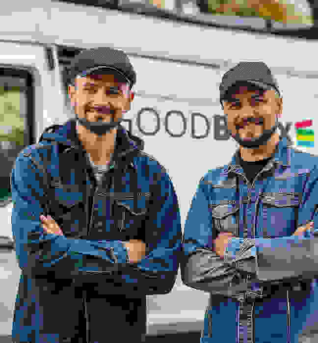 Zwei Männer in Jeansjacken und Mütze stehen mit verschränkten Armen vor einem weißen Van und lächeln.
