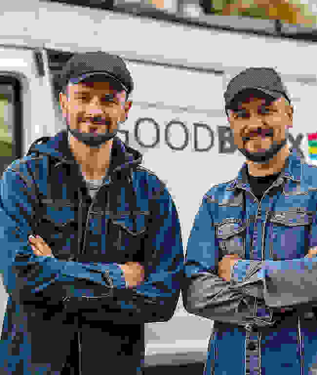 Zwei Männer in Jeansjacken und Mütze stehen mit verschränkten Armen vor einem weißen Van und lächeln.