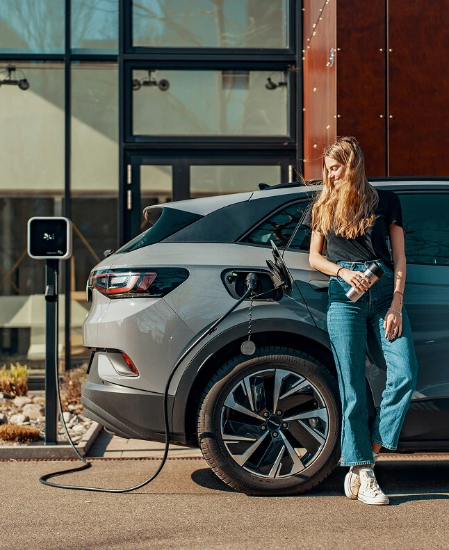 Eine junge Frau mit bräunlichem Haar lehnt lächelnd an einem grauen E-Auto, das an einer Ladestation angeschlossen ist.