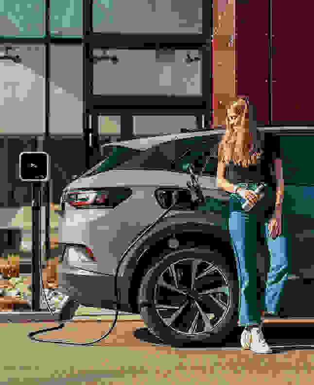 Eine junge Frau mit bräunlichem Haar lehnt lächelnd an einem grauen E-Auto, das an einer Ladestation angeschlossen ist.