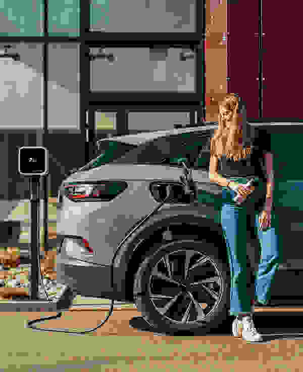 Eine junge Frau mit bräunlichem Haar lehnt lächelnd an einem grauen E-Auto, das an einer Ladestation angeschlossen ist.