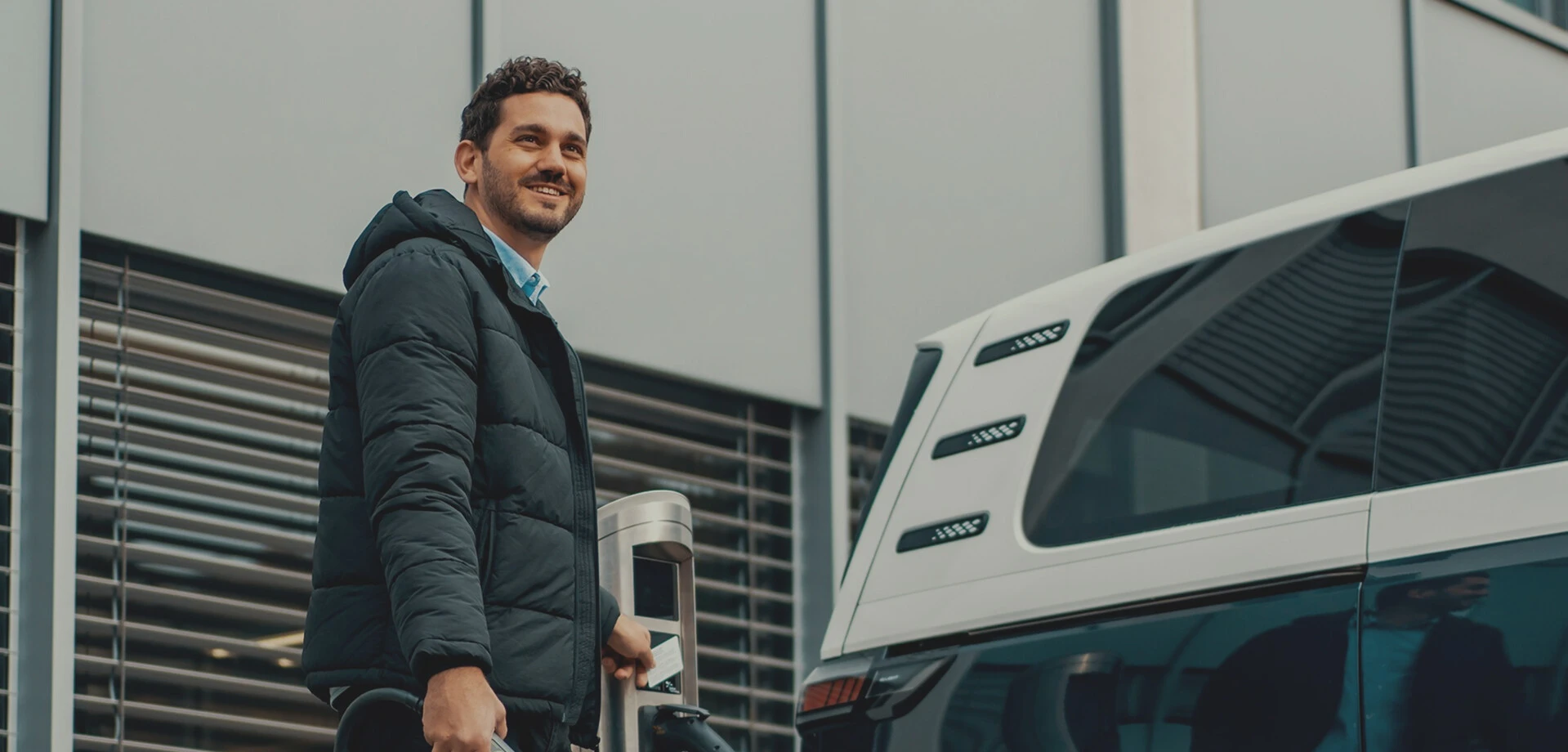 Ein junger Mann mit leicht lockigem braunem Haar und grauer Jacke blickt in die Ferne, während er neben einem weißen Auto steht und eine Karte an ein Gerät hält.