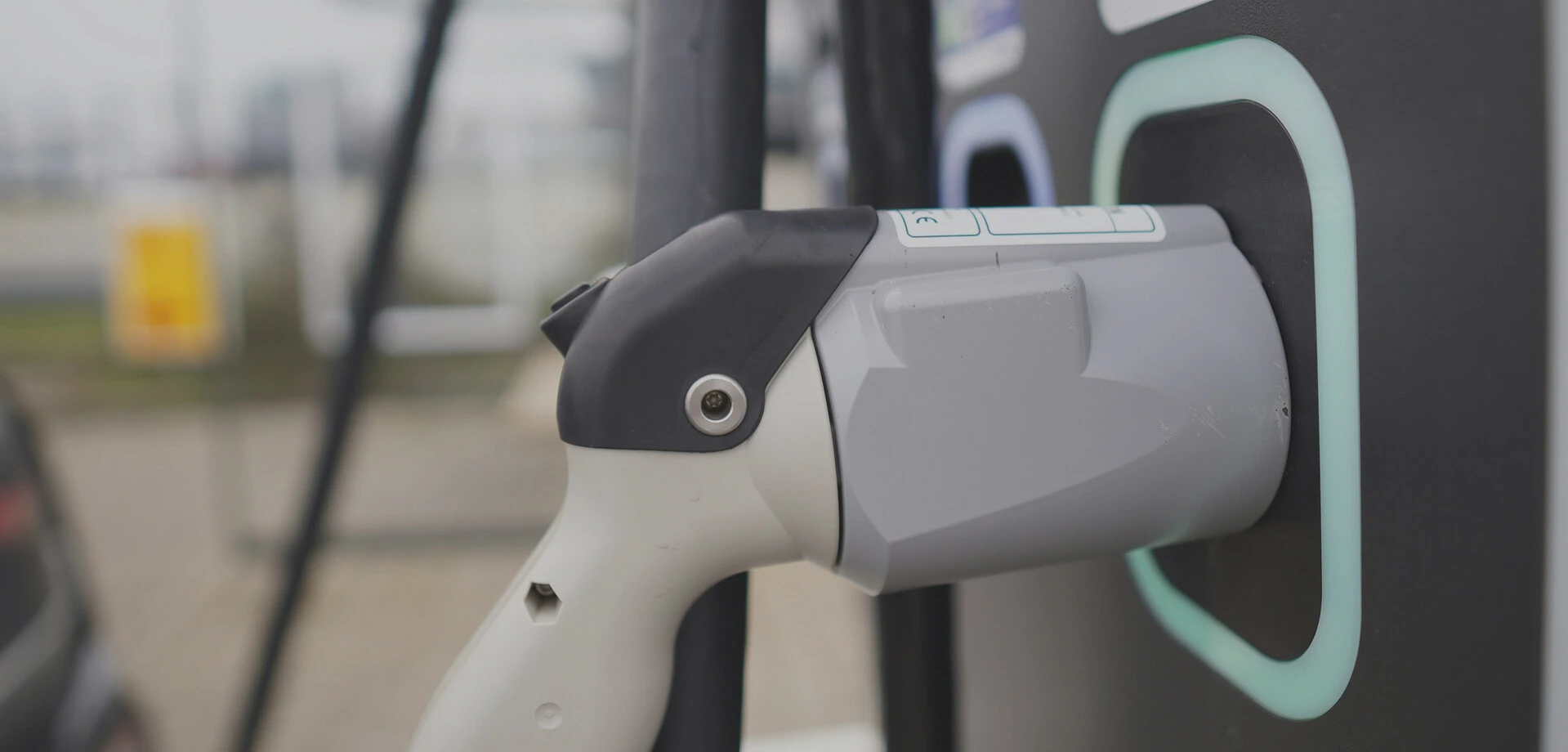 Ein weißes Ladegerät für E-Autos ist an einer grauen Ladestation mit mintfarbener Markierung befestigt – der Hintergrund ist unscharf.