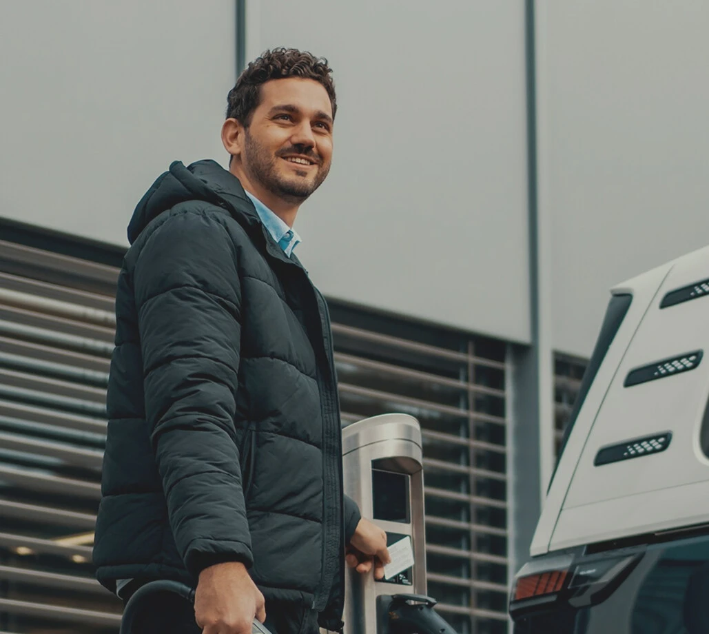 Ein junger Mann mit leicht lockigem braunem Haar und grauer Jacke blickt in die Ferne, während er neben einem weißen Auto steht und eine Karte an ein Gerät hält.